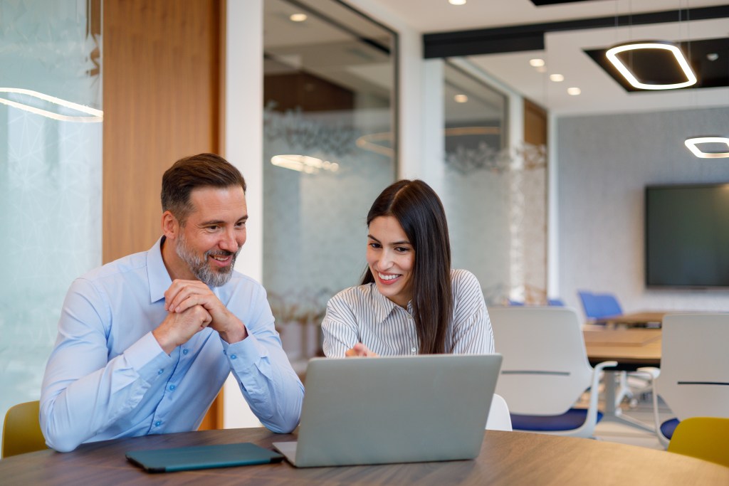 Man and woman smiling while looking at a computer in a modern office conference room.
